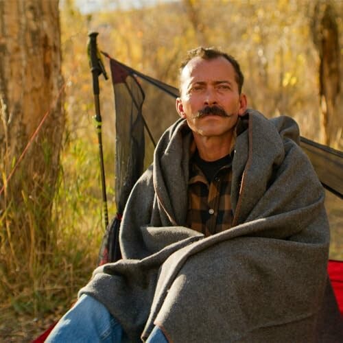 Man wrapped in blanket sitting in a campsite during autumn.