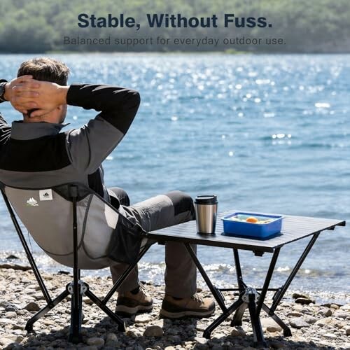 Man sitting on a lakeside in a camping chair with table.
